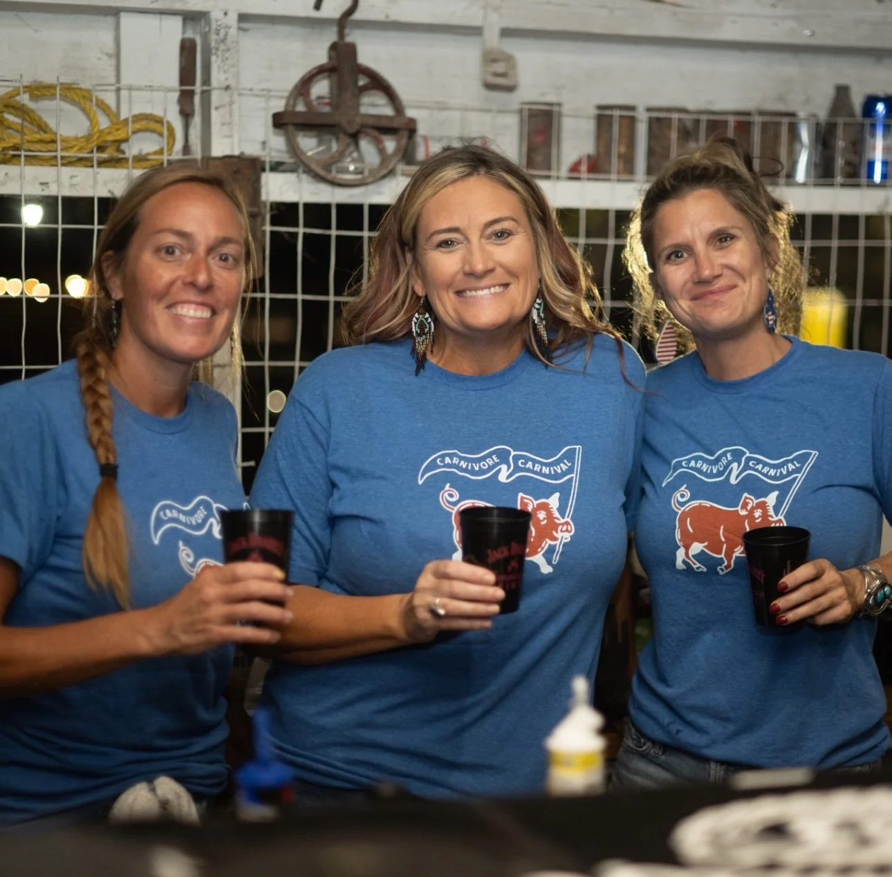 Three women wearing matching blue t-shirts with a pig and banner design, smiling and holding black cups at an outdoor night event.