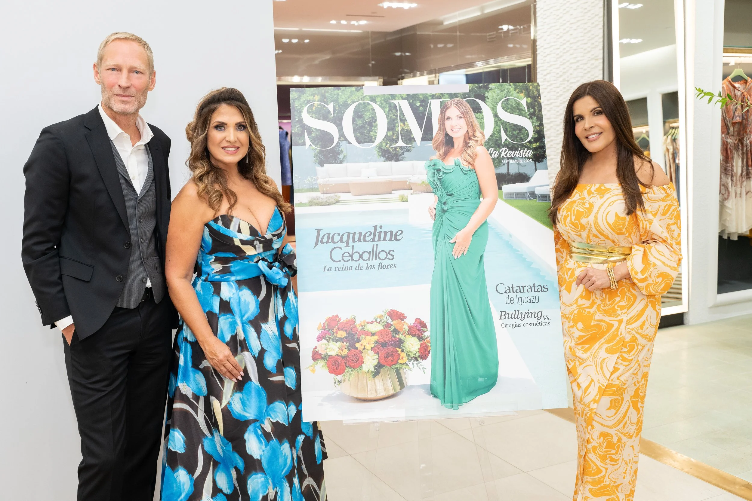 Four people standing indoors next to a magazine cover display, with one woman in a blue floral dress, a woman in an orange patterned dress, a man in a black suit, and another woman in a green dress. The display features the magazine title 'SÓVOS' and an image of a woman in a green gown by a pool.