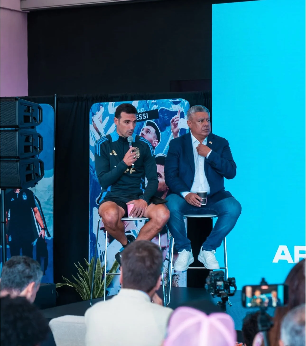 Two men on a panel discussion, one wearing a dark sports jacket and shorts, holding a microphone, and the other in a blazer and jeans, holding a cup. They are seated on stools in front of a blue background with a poster of a soccer player.