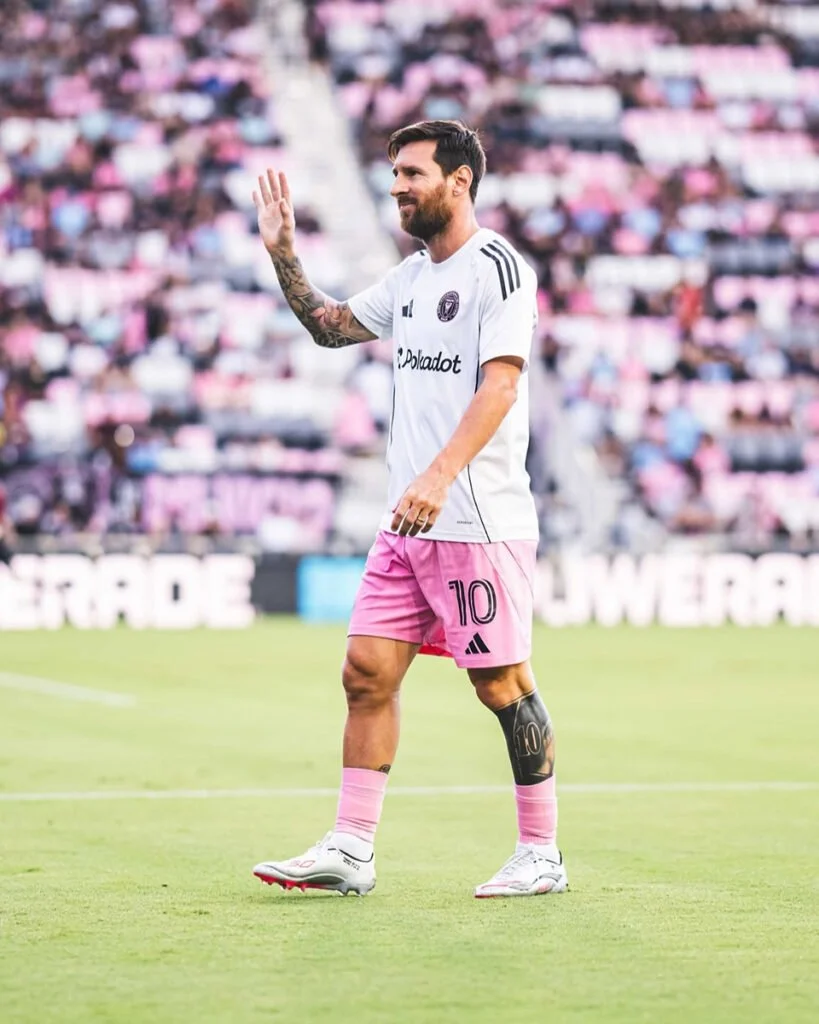 A soccer player wearing a white jersey and pink shorts, standing on the field and waving to the crowd during a match.
