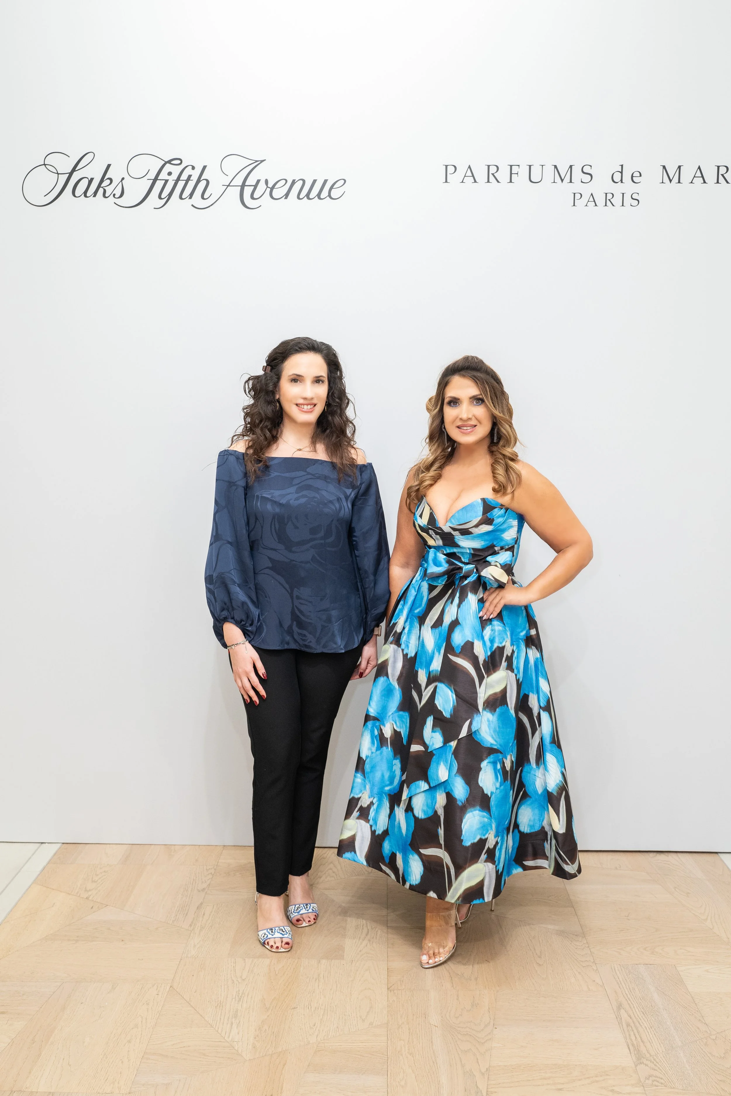 Two women in elegant dresses posing in front of a white wall with black script text that reads "Saks Fifth Avenue" and "Parfums de Marly Paris."