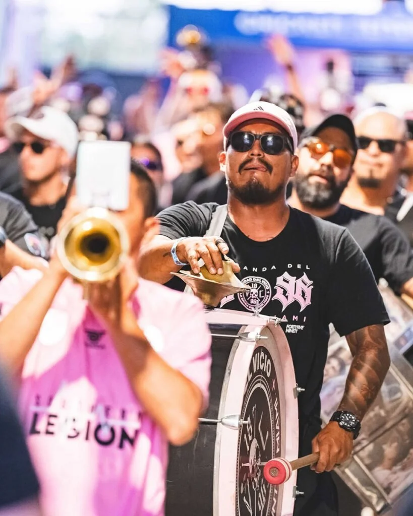 A man playing drum with a percussion instrument at a lively event surrounded by a crowd, including a trumpet player in a pink shirt in the foreground.