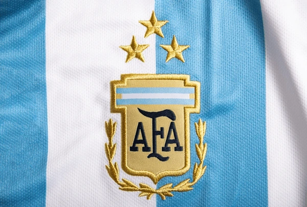 Close-up of the Argentine national football team emblem embroidered on a jersey, featuring three gold stars above the shield, blue and white vertical stripes background.