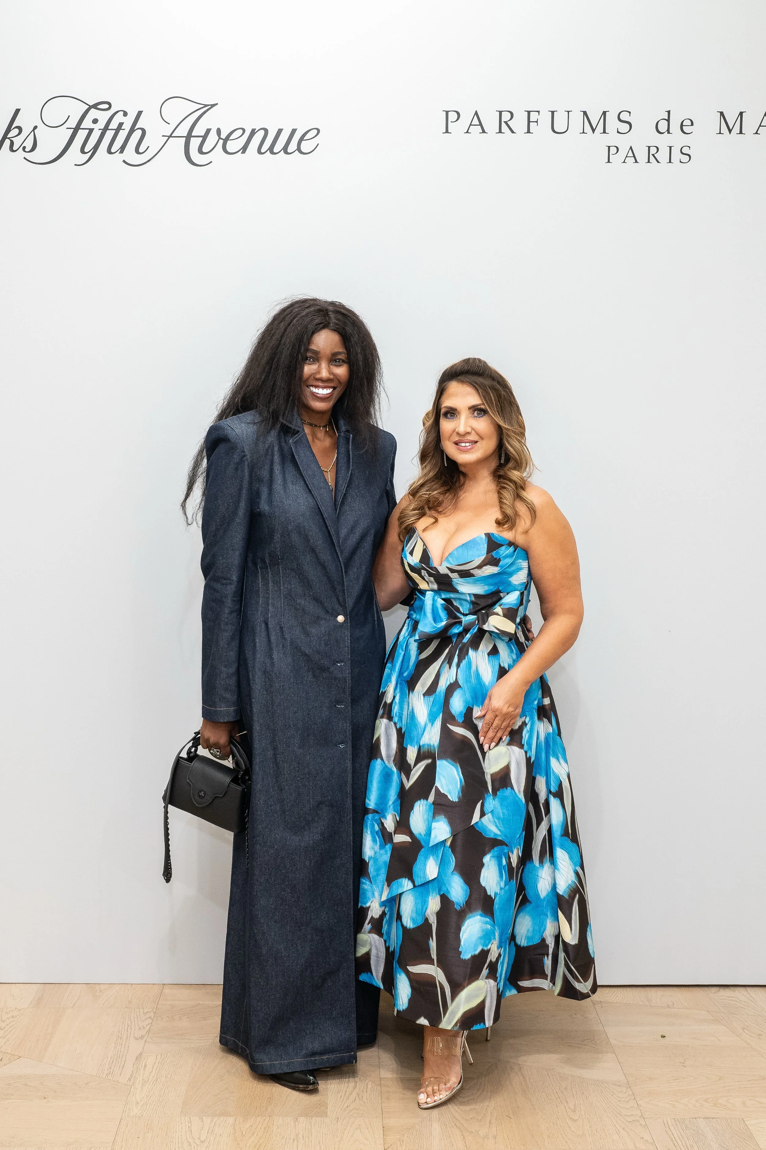 Two women standing together, smiling, in front of a white wall with the text 'Thanks Fifth Avenue' and 'PARFUMS de MARQUES PARIS'. One woman is dressed in a dark denim outfit holding a black purse, and the other is wearing a blue and black floral dre