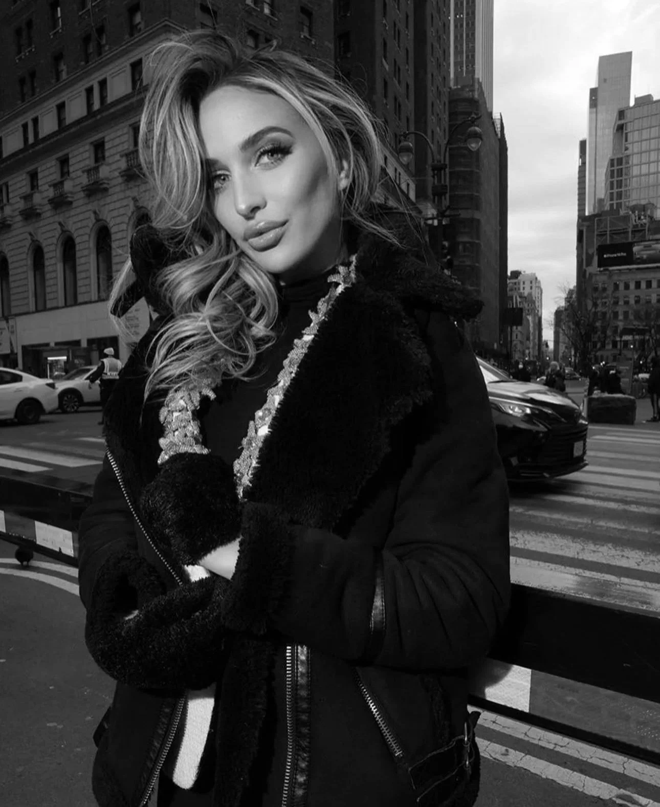 Black-and-white photo of a woman with wavy hair in a city street, leaning on a railing, wearing a dark coat and gloves.