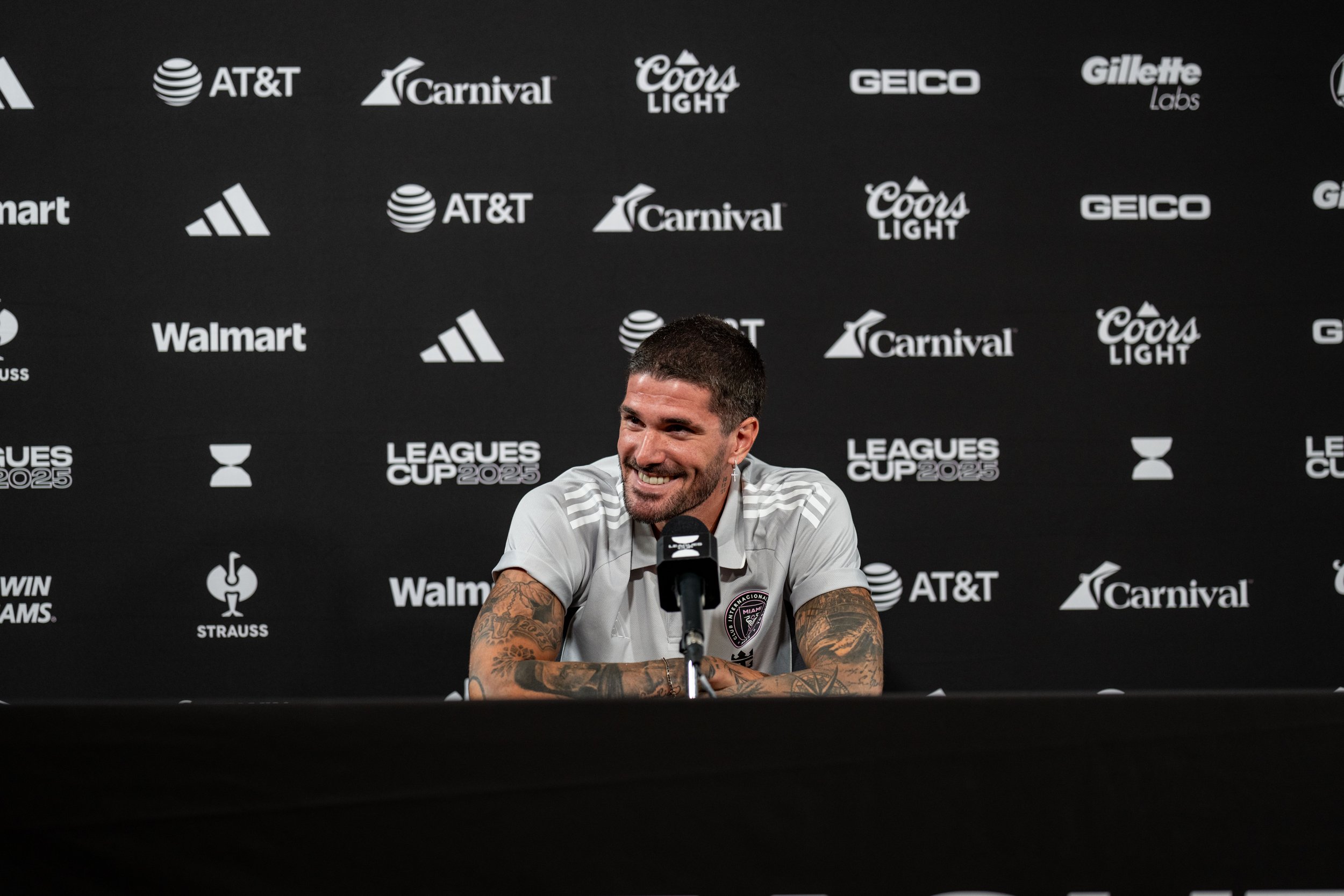 A man with tattoos sitting at a press conference table, smiling, with a black backdrop featuring various sponsor logos, including AT&T, Carnival, Coors Light, GEICO, Gillette Labs, Walmart, Adidas, and Strauss, at the Leagues Cup 2025 event.