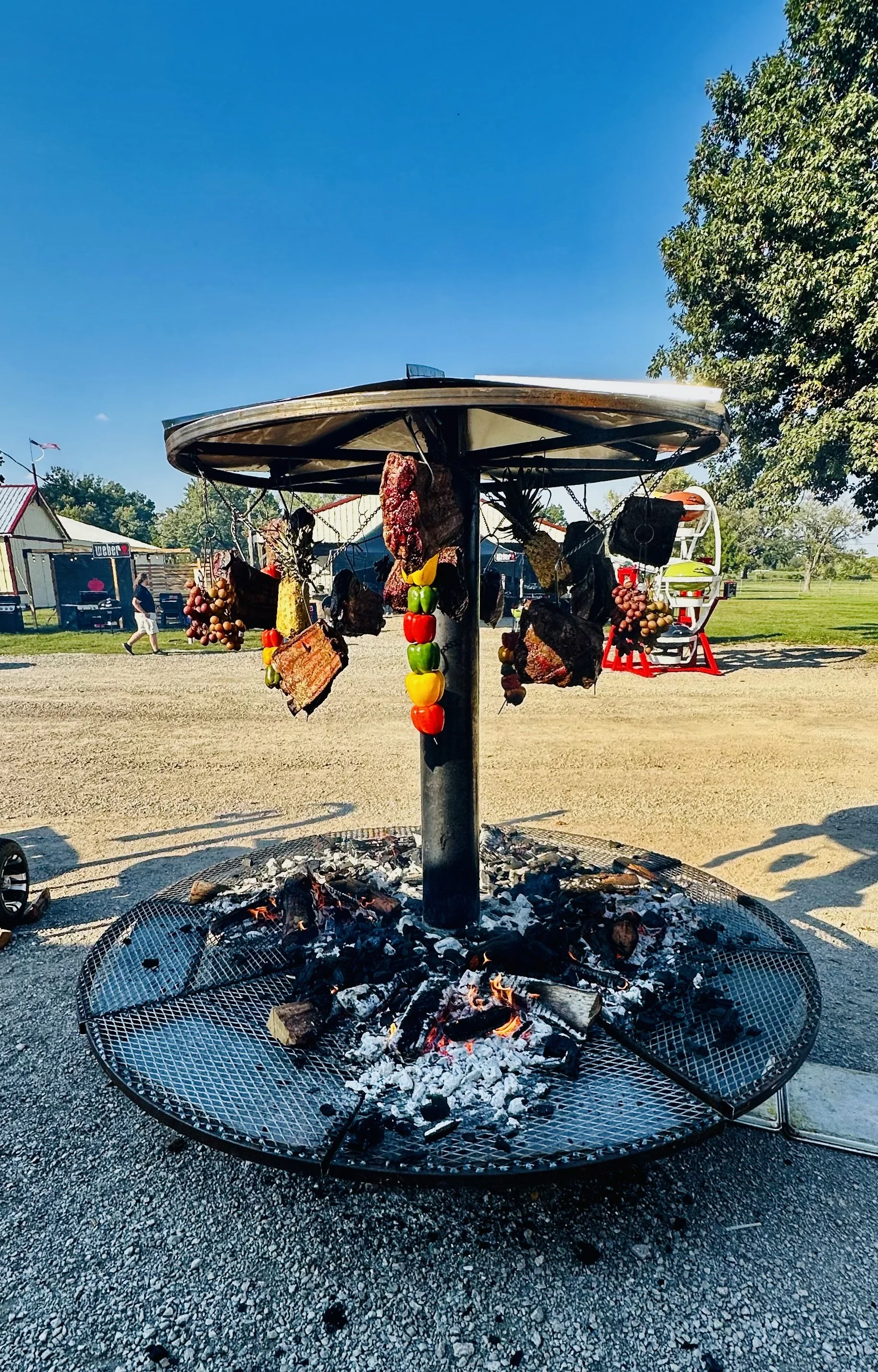 A cowboy-style spit roasting pit with hanging meat and colorful peppers, set outdoors on a sunny day.