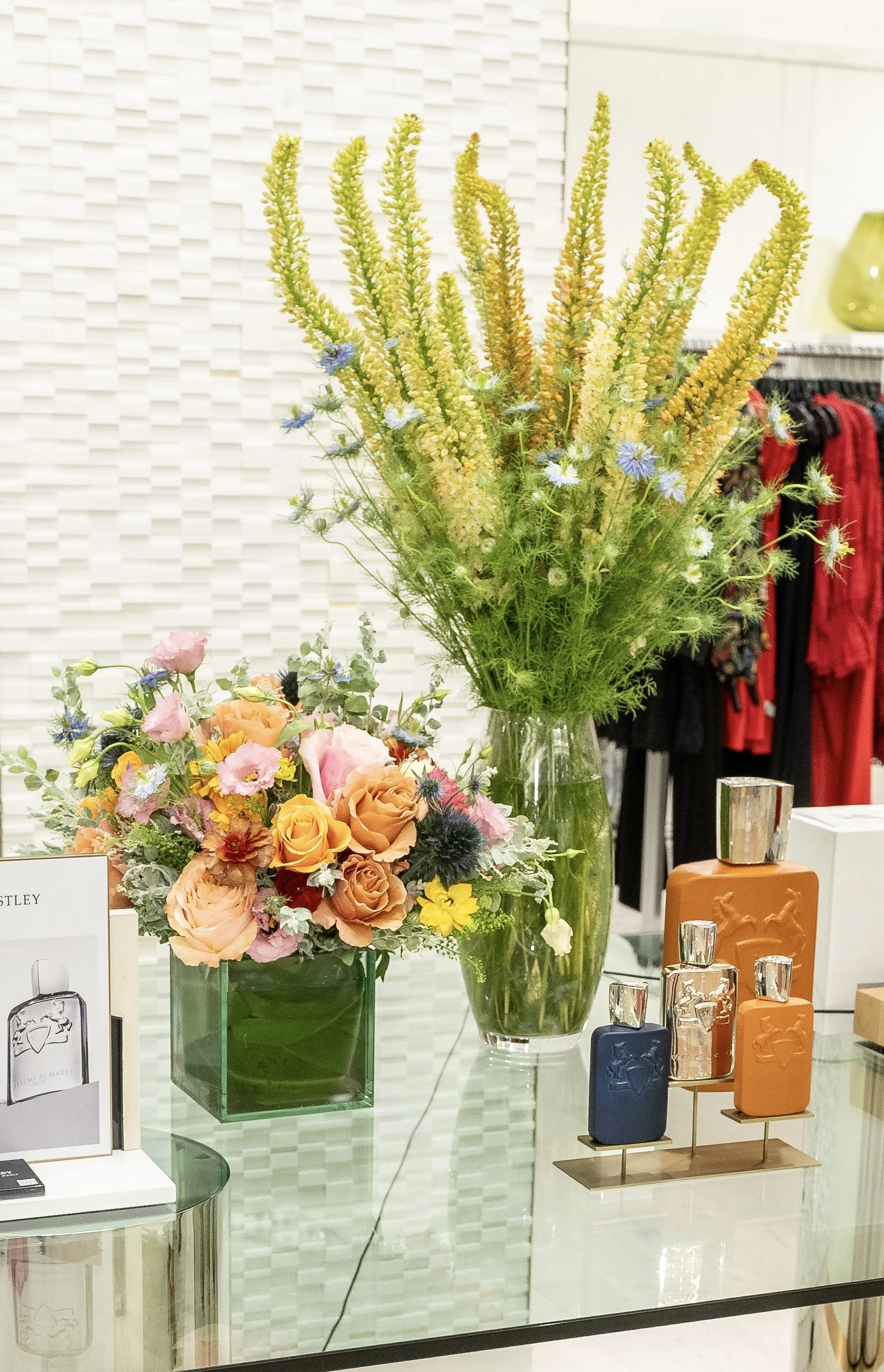 Display of vases with colorful flowers and perfume bottles on a glass table in a retail store.