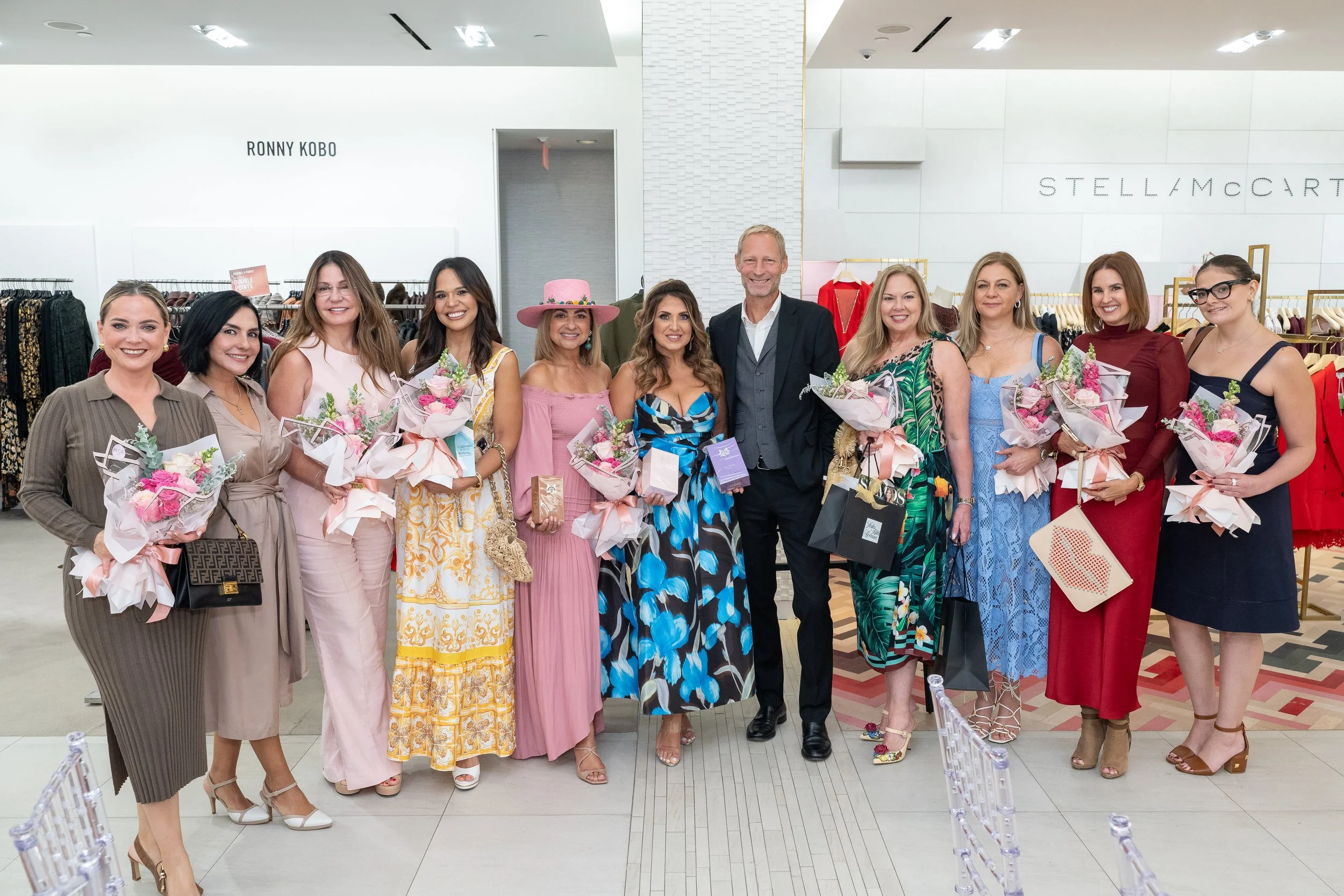 Group of women and one man standing indoors, holding floral bouquets and shopping bags, dressed in colorful attire, with clothing racks and store signage in the background.