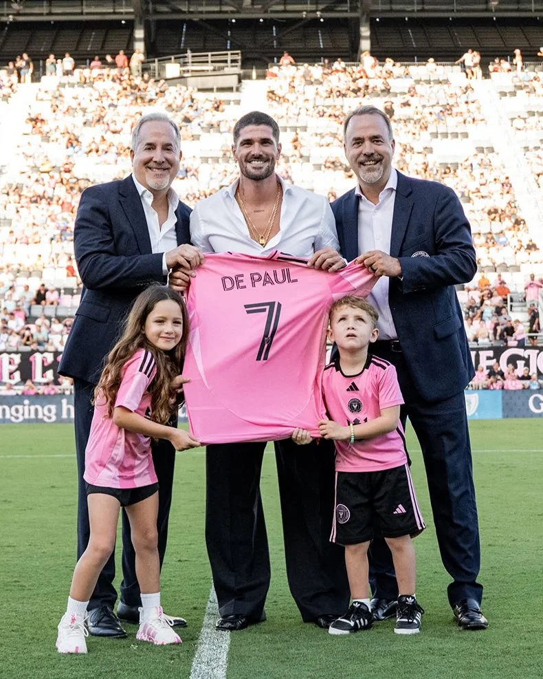 Group of five people on a soccer field, holding up a pink soccer jersey with the name 'De Paul' and number '7'. The group includes a man in a suit, a man in a white shirt, a man in a blazer, and two children in pink soccer jerseys. The stadium is full of spectators.