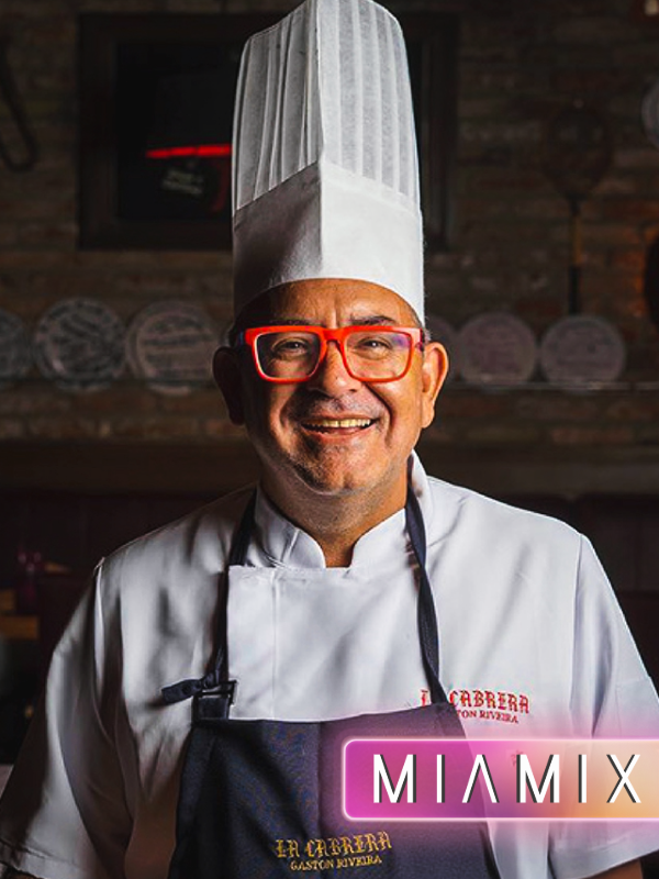 Smiling male chef wearing a white chef's hat and coat with red glasses, black apron with yellow text, in a rustic restaurant setting.