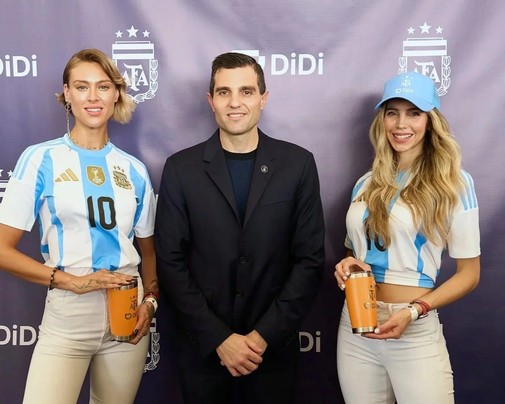 Two women in Argentina soccer jerseys holding drink tumblers, standing next to a man in a black jacket, in front of a backdrop with Argentina Football Association logos and the word 'Didi'.