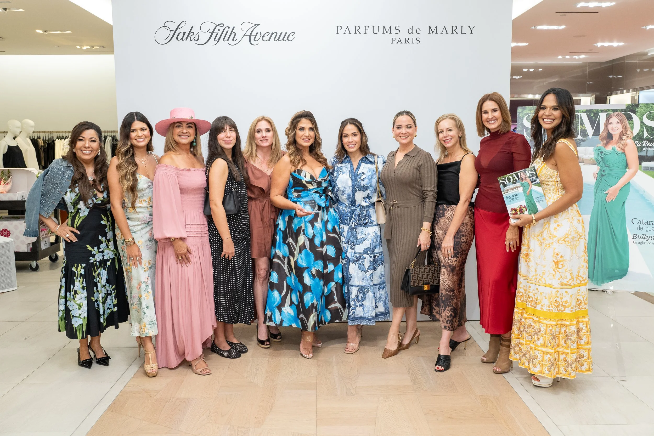 Group of women standing together inside a store, smiling, with a sign behind them that reads 'Saks Fifth Avenue' and 'Parfums de Marly Paris'.