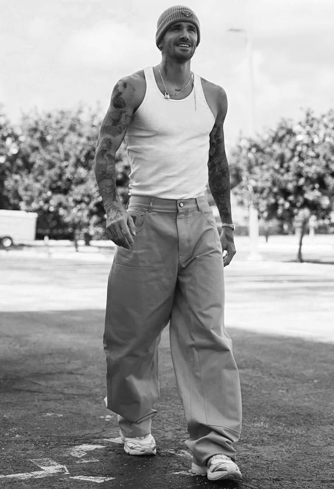 A man walking outdoors in casual clothing, wearing a beanie hat, tank top, loose pants, sneakers, and tattoos visible on his arms, smiling.