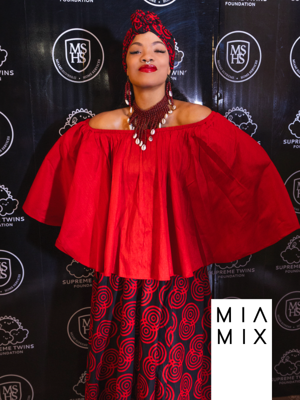 A woman in a red off-shoulder top with loose, flowing sleeves, featuring bold patterned pants with red swirls and circles, standing in front of a black backdrop with white logos and text, wearing a red and black patterned headwrap, red lipstick, and statement jewelry including a necklace and earrings.