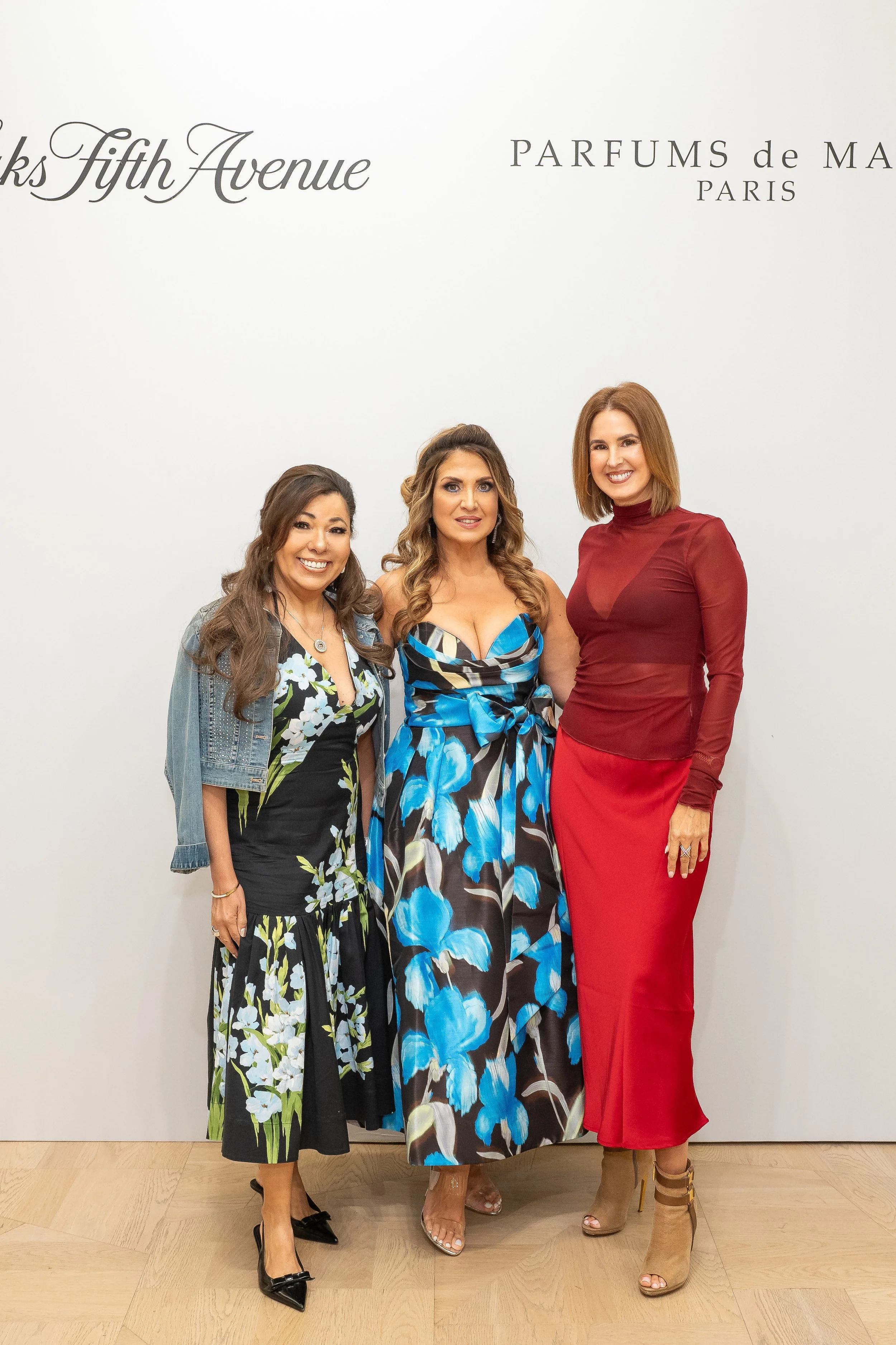 Three women standing together in front of a white wall with black text that reads 'Fifth Avenue' and 'PARFUMS de MA PARIS.' They are dressed in fashionable dresses, smiling, and posing for the photo.