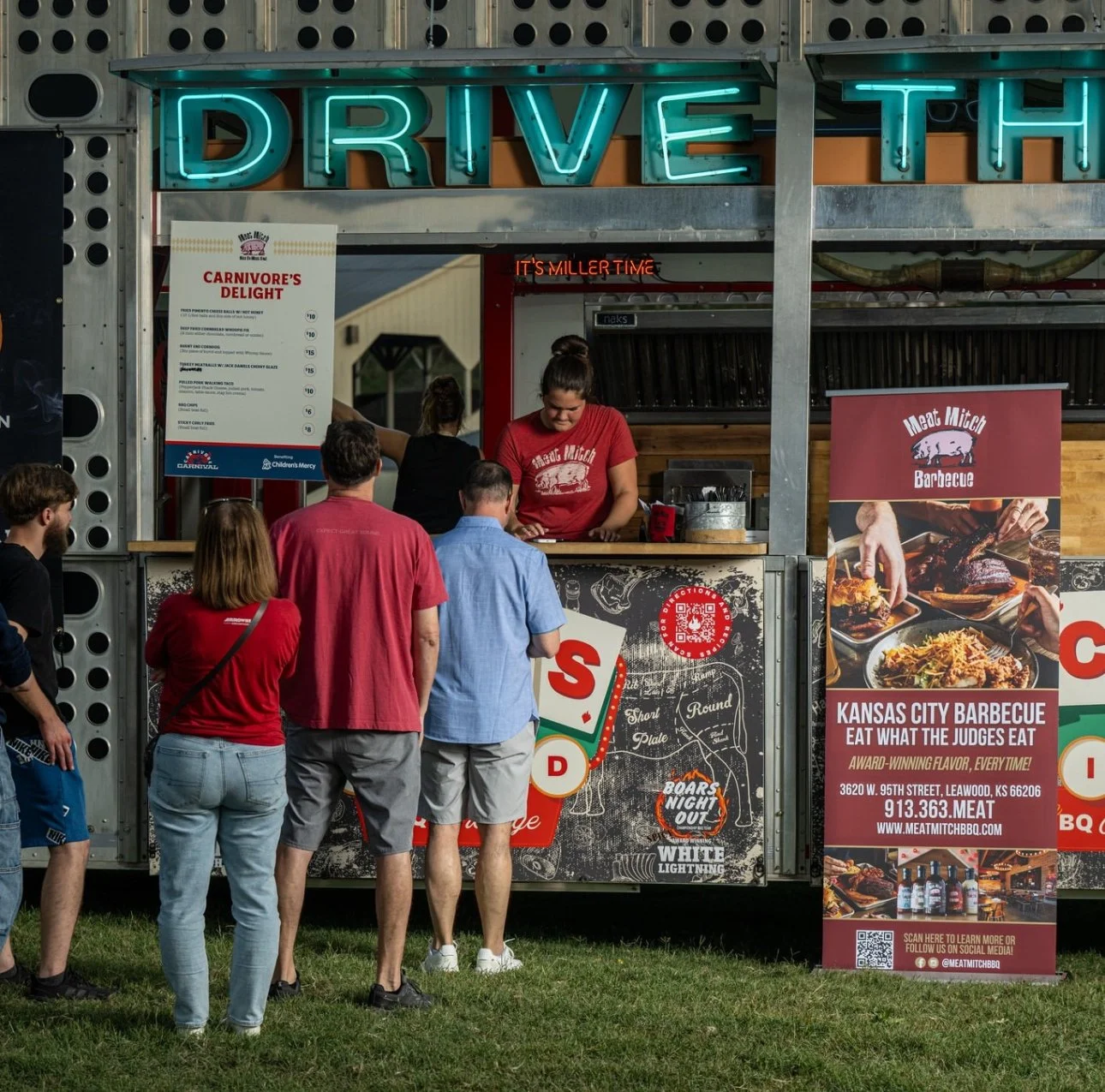 People ordering barbecue food at Kansas City barbecue stand with neon 'Drive Thru' sign, menu, and promotional banner.