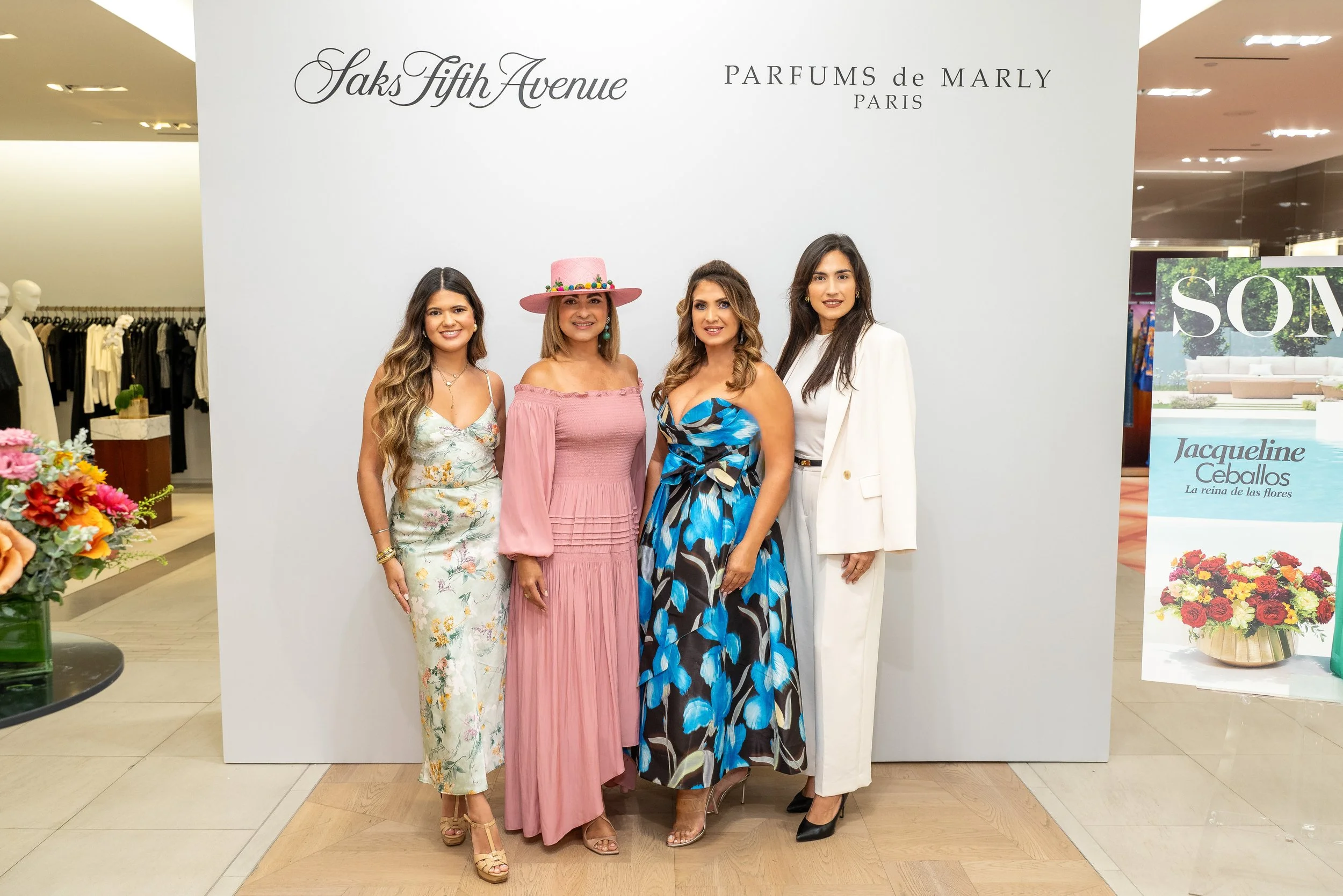 Four women in fashionable attire standing in front of a white backdrop with text. They are inside a shopping mall or department store, with a floral display and retail clothing visible around them. The backdrop features the text 'Jaks Fifth Avenue' a