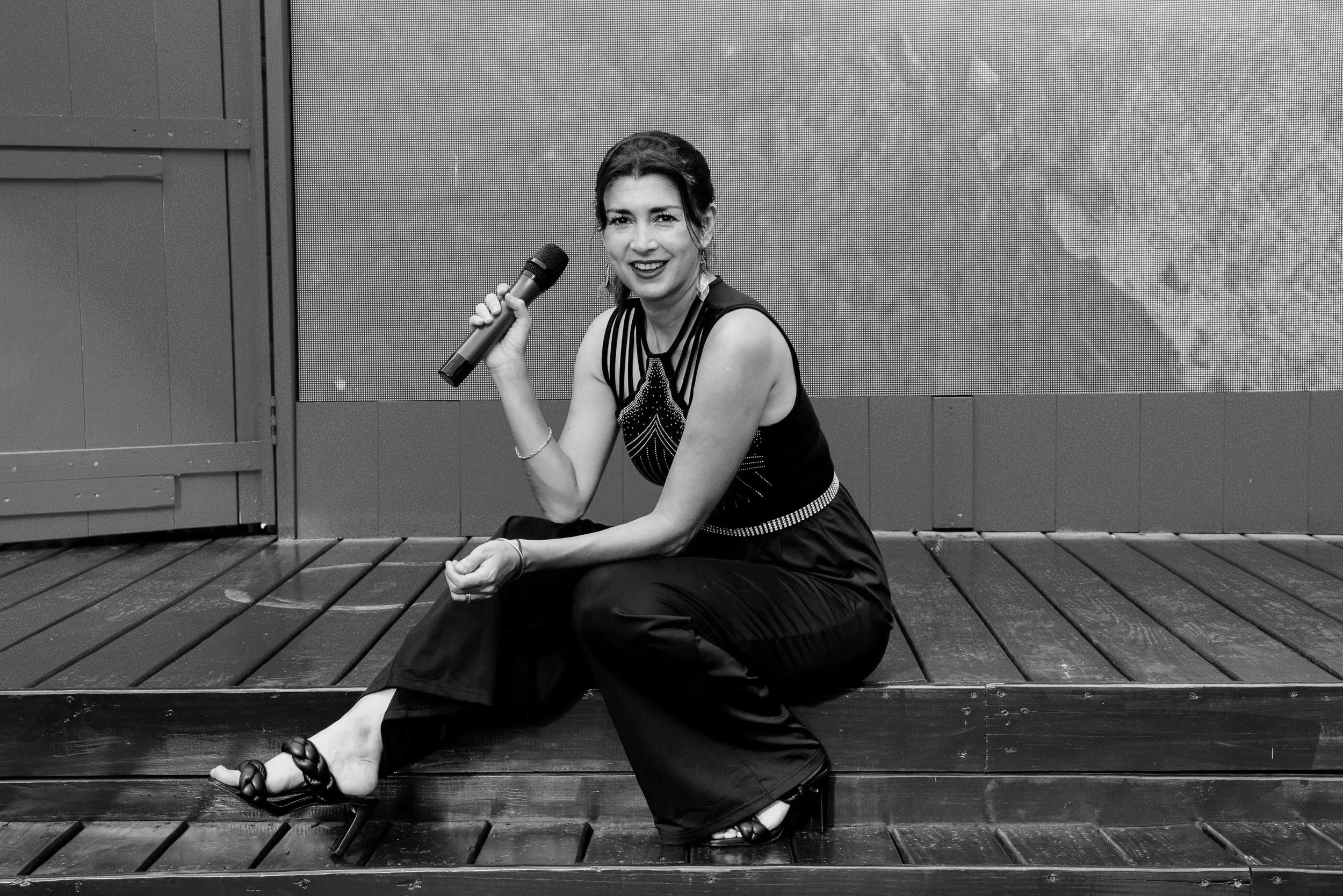 A woman sitting on a wooden platform, holding a microphone, smiling, dressed in a sleeveless top and wide-leg pants, with a large screen behind her.