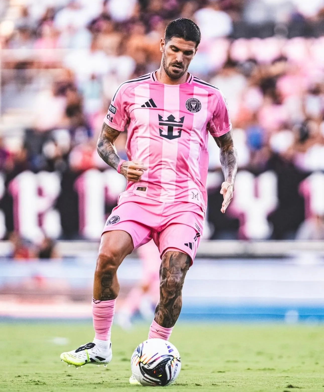 A male soccer player in a pink uniform standing on a soccer field, with a crowd in the background, preparing to kick a soccer ball.