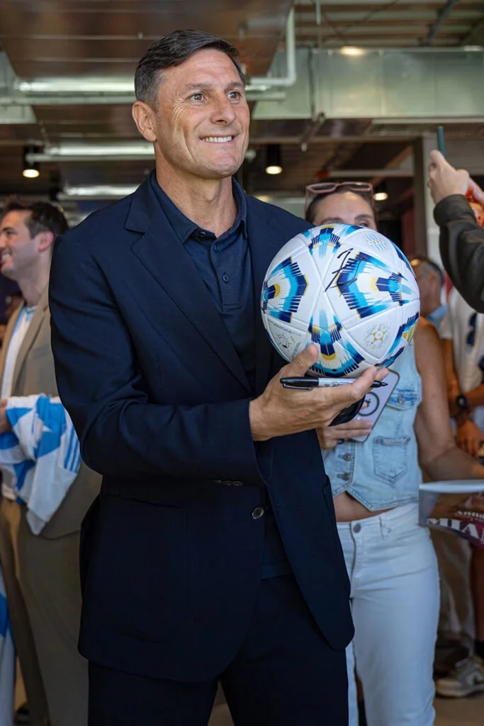 A man in a navy blazer holding a soccer ball decorated with colorful patterns at a public event, with people around him taking photos or looking on.