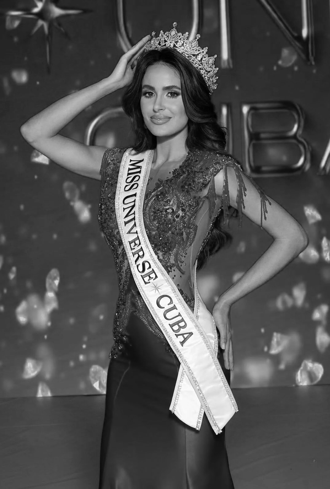 Miss Universe Cuba contestant wearing a crown and sash, posing with one hand on her hip against a decorated backdrop.