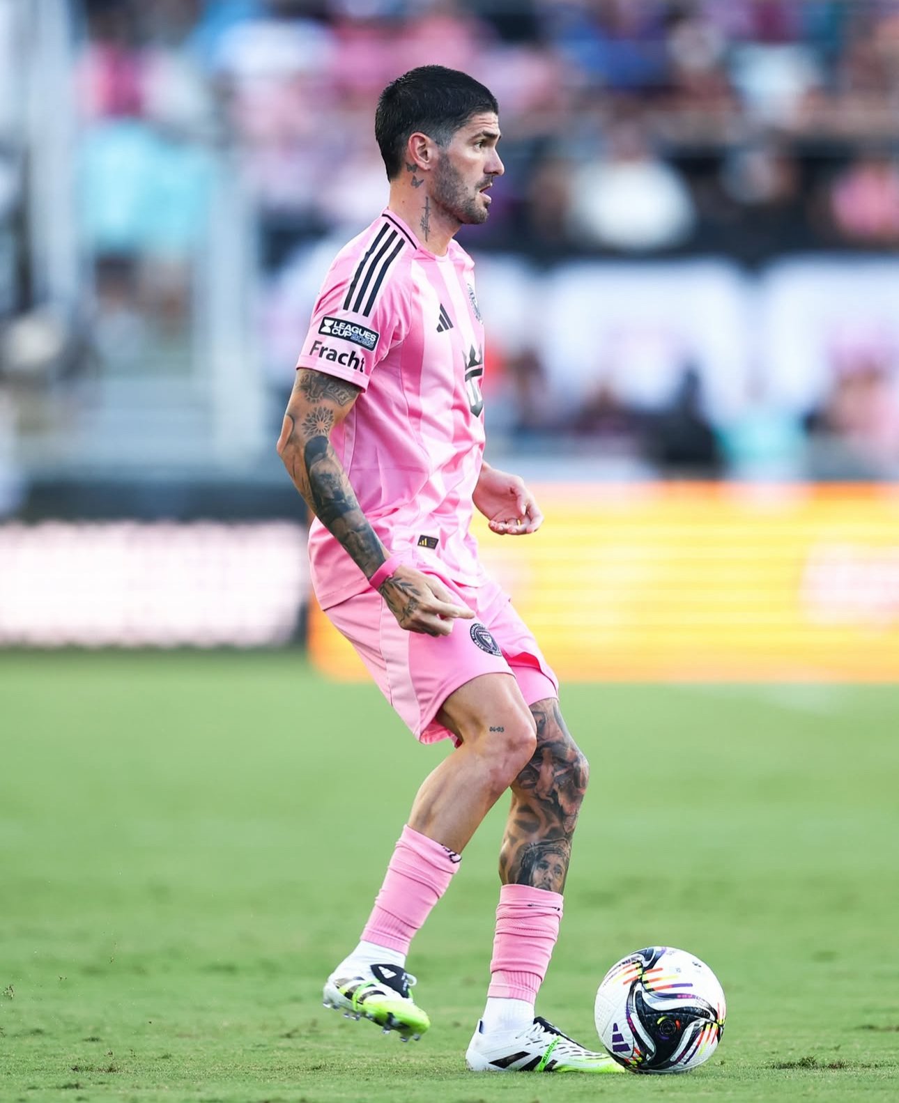 A male soccer player with tattoos on his arms and legs, wearing a pink jersey and pink shorts, standing on a soccer field while controlling a soccer ball with his foot.