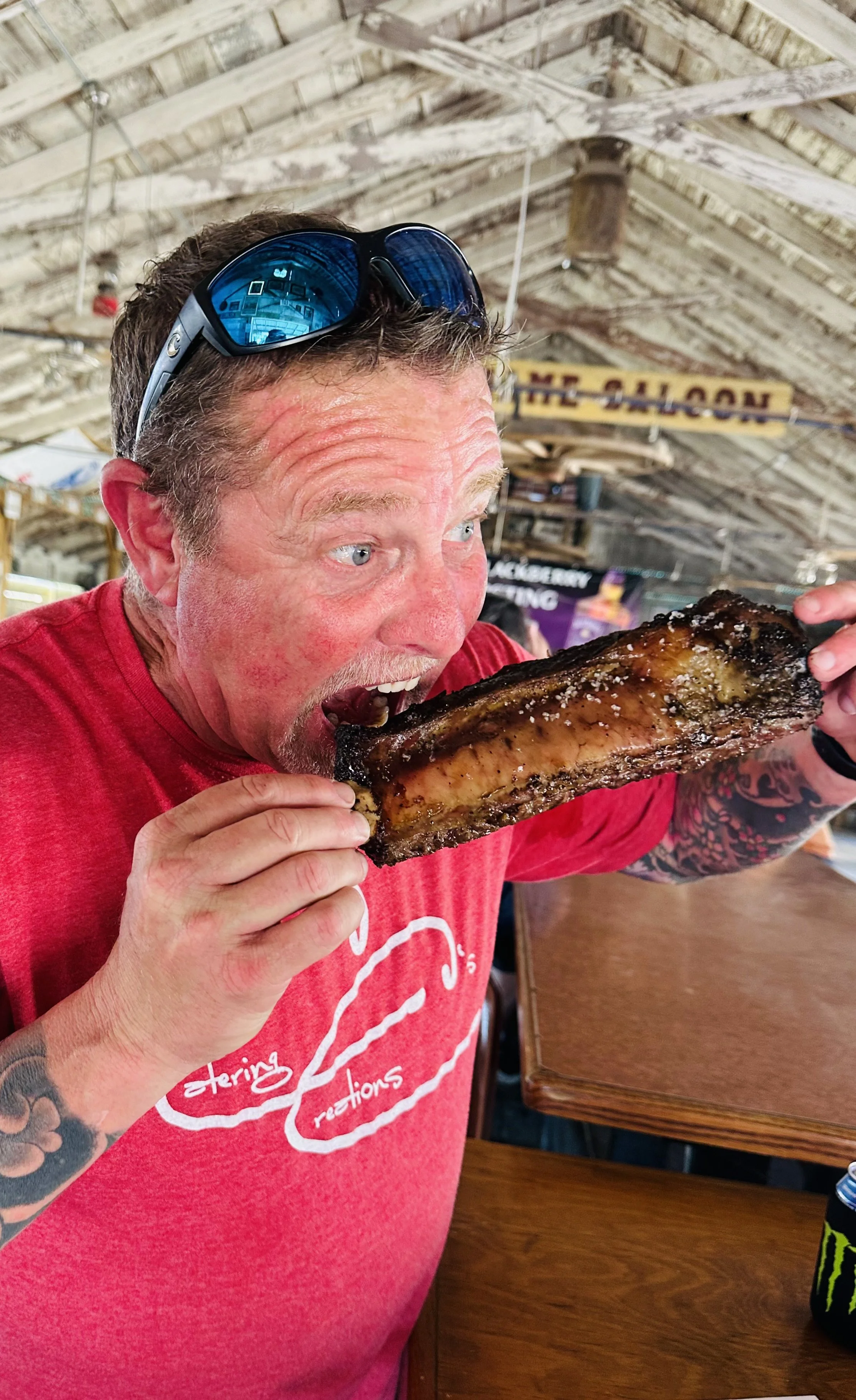 A man with tattoos on his arm wearing a red t-shirt with a burger icon and text, sunglasses on his head, about to bite into a large piece of cooked ribs at an indoor restaurant with a rustic, wooden roof in the background.