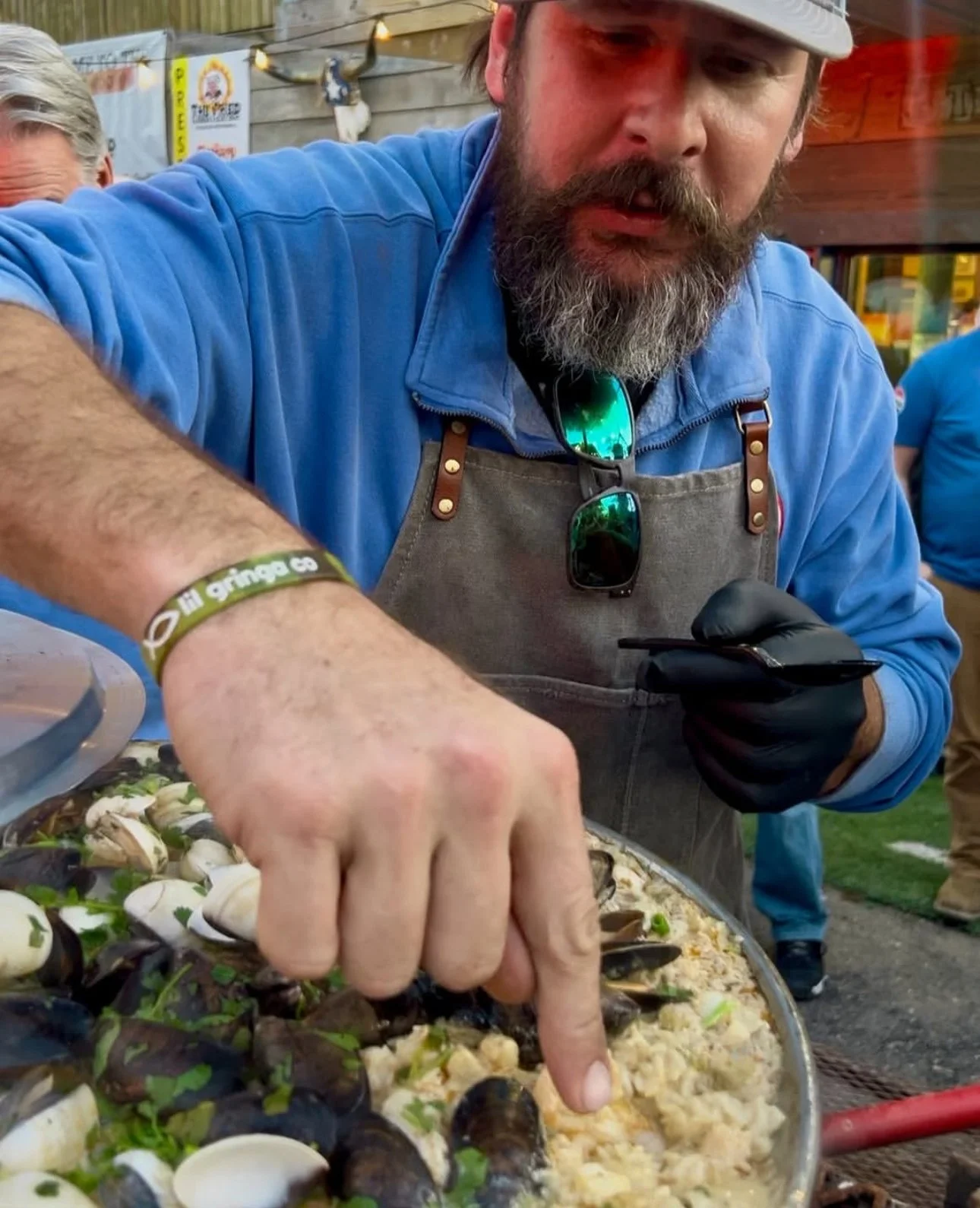 A man with a beard wearing a gray apron and black gloves is preparing a seafood dish, garnished with herbs, at an outdoor event.