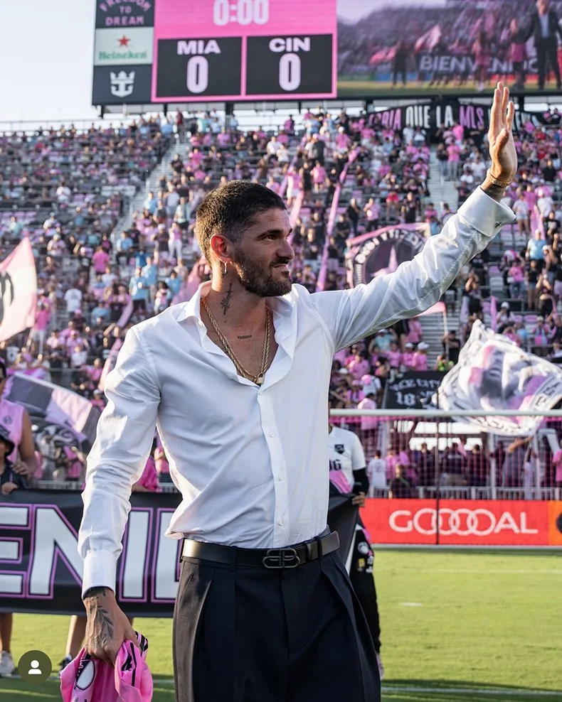 A man with a beard wearing a white shirt and black pants waves to the crowd at a football stadium. Fans in the stands wear pink and black, and banners are visible. The scoreboard shows a score of 0-0 between Miami and Cincinnati.