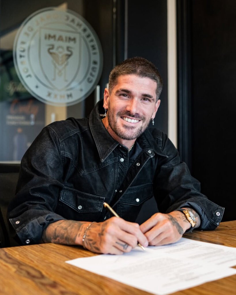 A smiling man with short dark hair and beard, wearing a black denim jacket, sitting at a wooden table, holding a pen over a document, with a glass of water nearby and a window with a logo in the background.