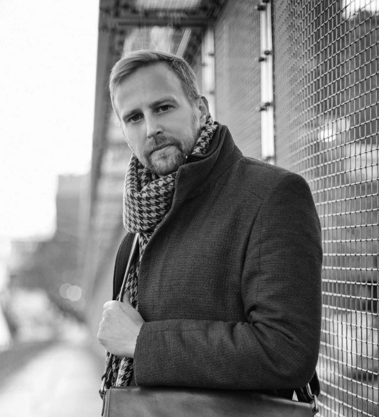 A black and white photo of a man with a beard and short hair wearing a coat and a patterned scarf, standing against a chain-link fence.