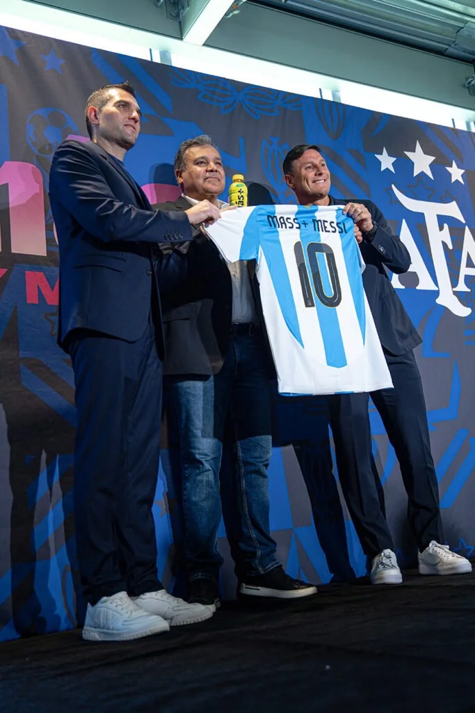 Three men on stage holding a light blue and white soccer jersey with the name 'Messi' and the number 10, with a colorful background featuring stars and football motifs.