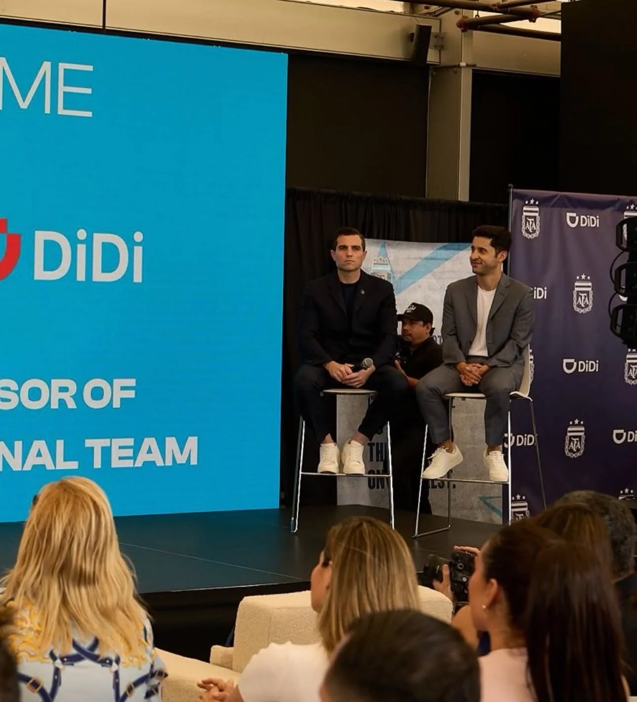 Two men sitting on high chairs on stage during a panel discussion, with an audience watching. The stage has a large blue screen with the logo 'Didi' and text mentioning 'Advisor of National Team.' The backdrop has multiple 'Didi' logos and a crest, p