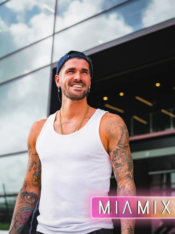 A smiling man with tattoos wearing a white tank top, black backwards baseball cap, and gold chains, standing outside a modern glass building.