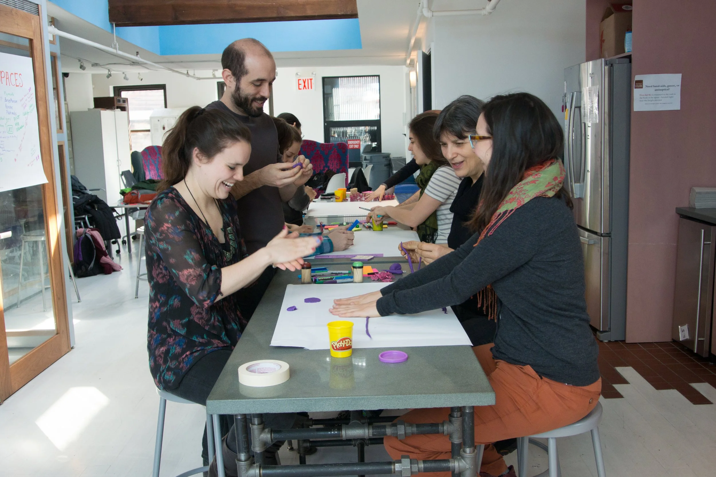 People gathered around a table in a workshop or classroom, engaged in arts and crafts activities with Play-Doh and colored paper.