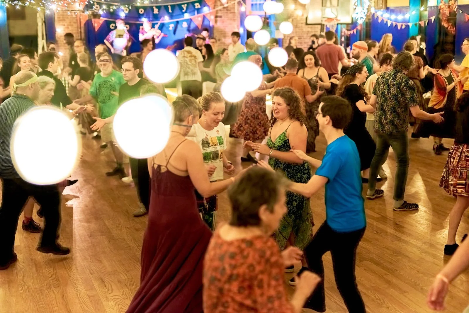 People dancing at a lively party with string lights, hanging paper lanterns, and a band on stage in the background.