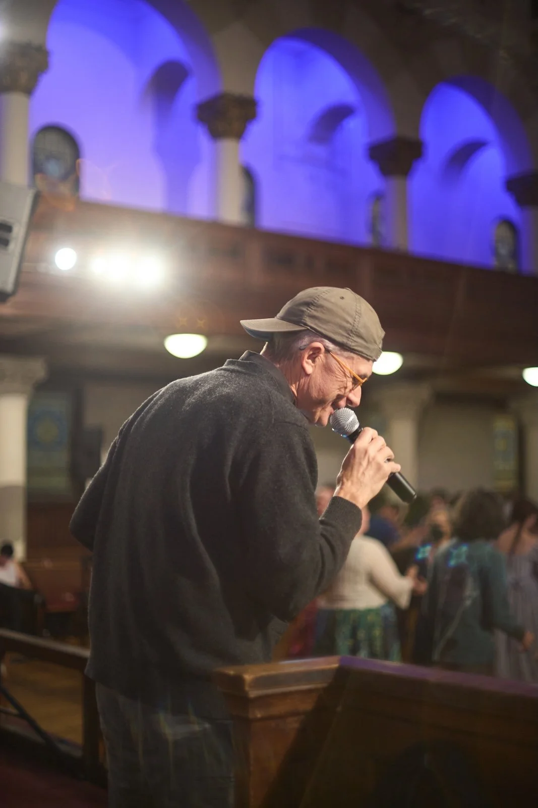 A man with gray hair wearing a beige cap and glasses, singing into a microphone inside a church or hall with blue lighting and arched windows.