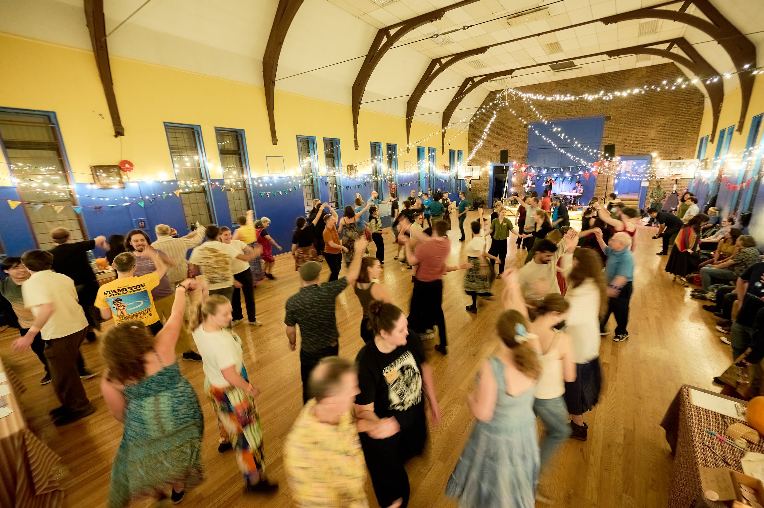 People dancing and socializing in a decorated event hall with string lights, banners, and a stage with a band playing.