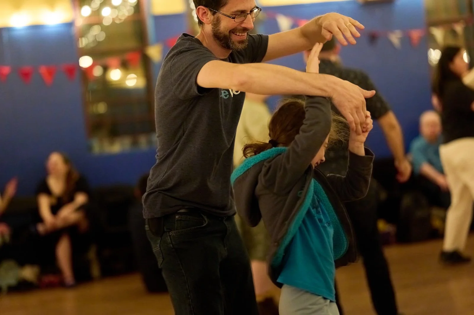 A man and a young girl dancing together at an indoor event, with other people seated in the background.