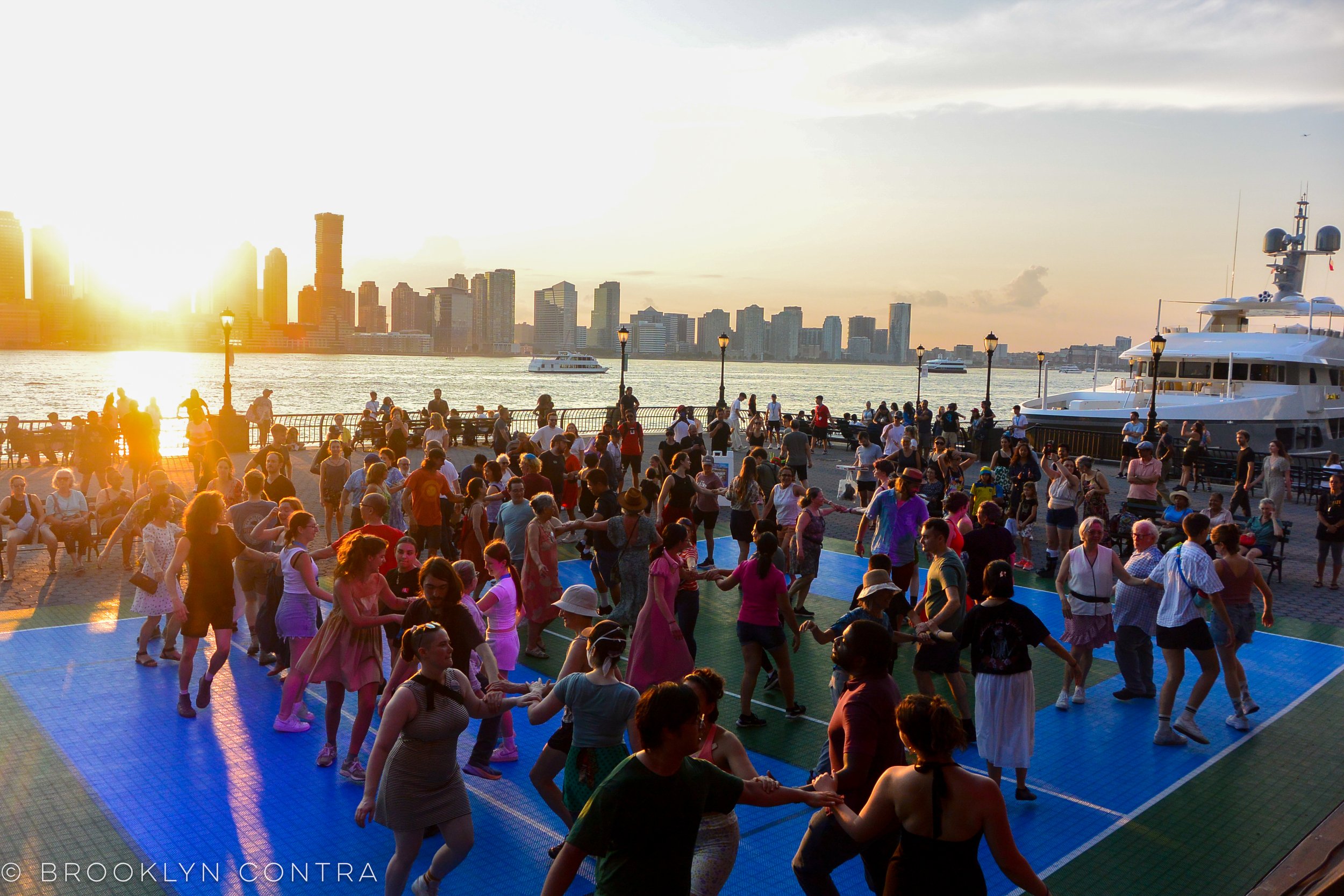 People dancing outdoors near a waterfront with a city skyline and boats in the background at sunset.