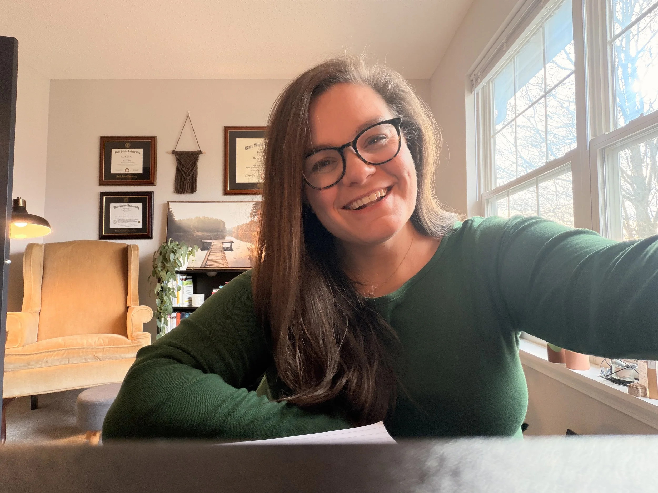 A woman with long brown hair, glasses, and a green sweater smiling while taking a selfie at a desk in a cozy home office. Behind her is a yellow armchair, framed certificates on the wall, a plant, and a bookshelf. Sunlight streams through large windows.