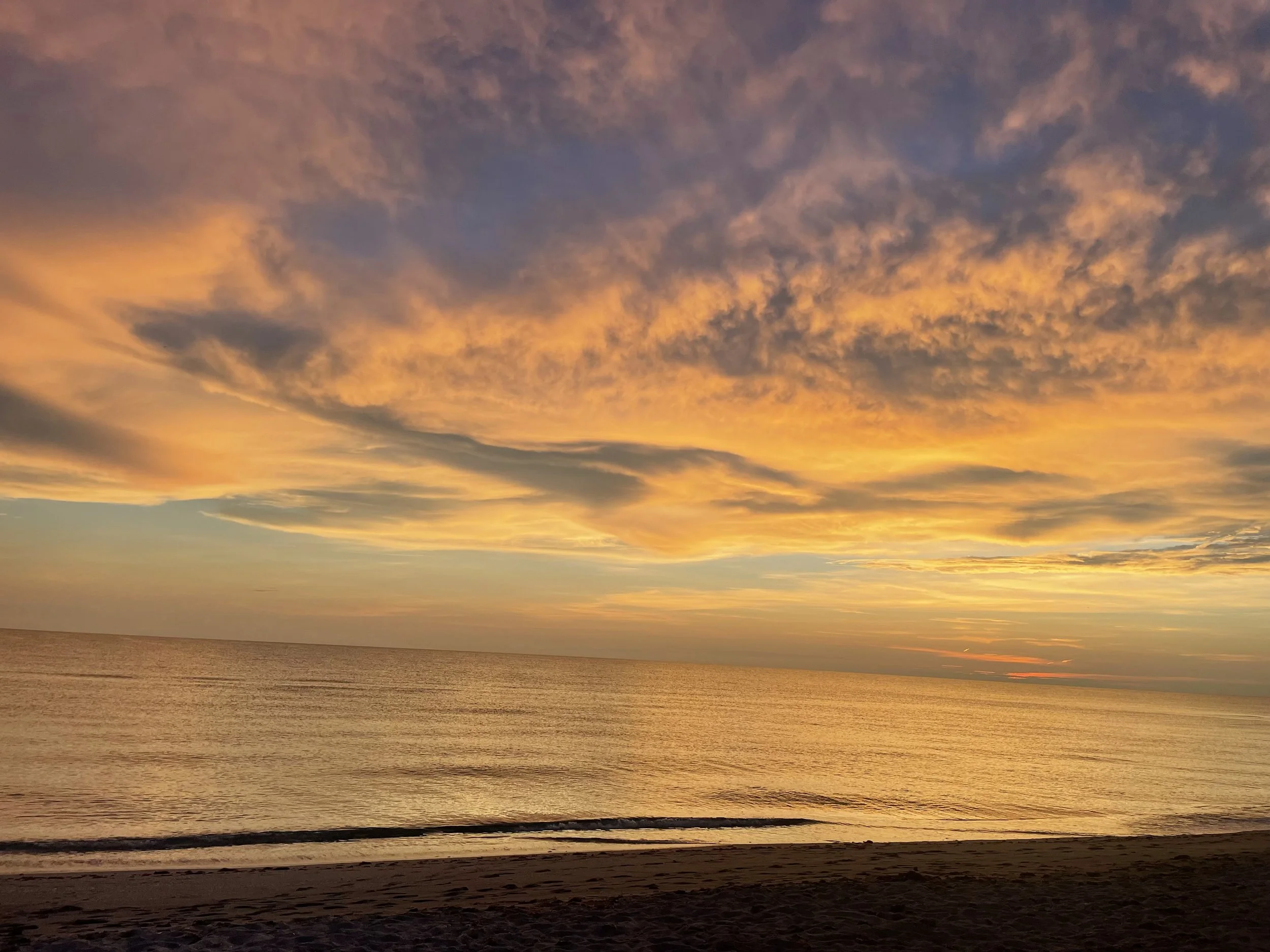Sunset over the ocean with orange and purple clouds and a sandy beach in the foreground.