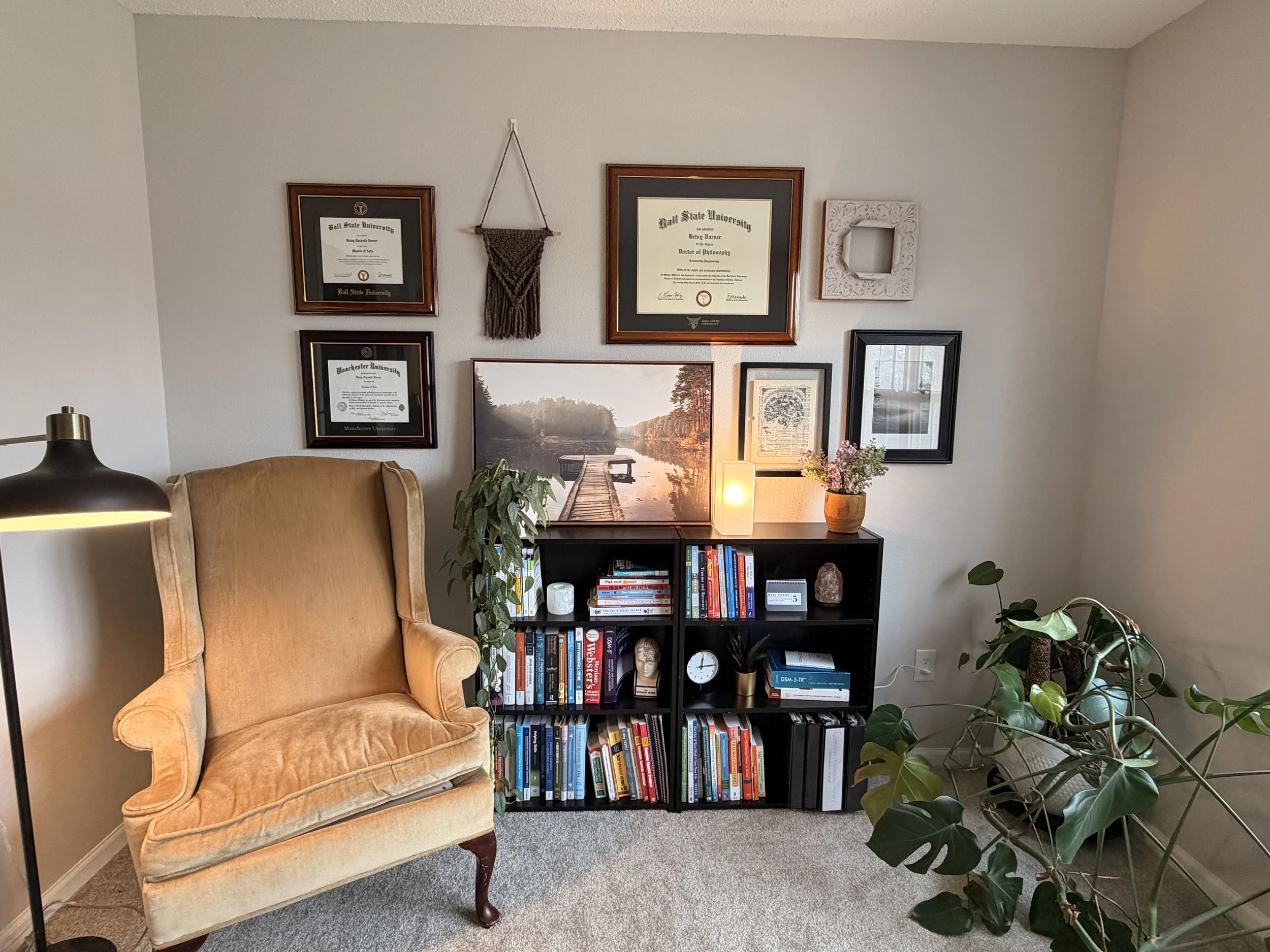 A cozy living room corner with a mustard-colored vintage armchair, black bookshelf filled with books and decor, framed certificates and artwork on the wall, and several indoor plants including a large Monstera and a potted plant on the shelf.