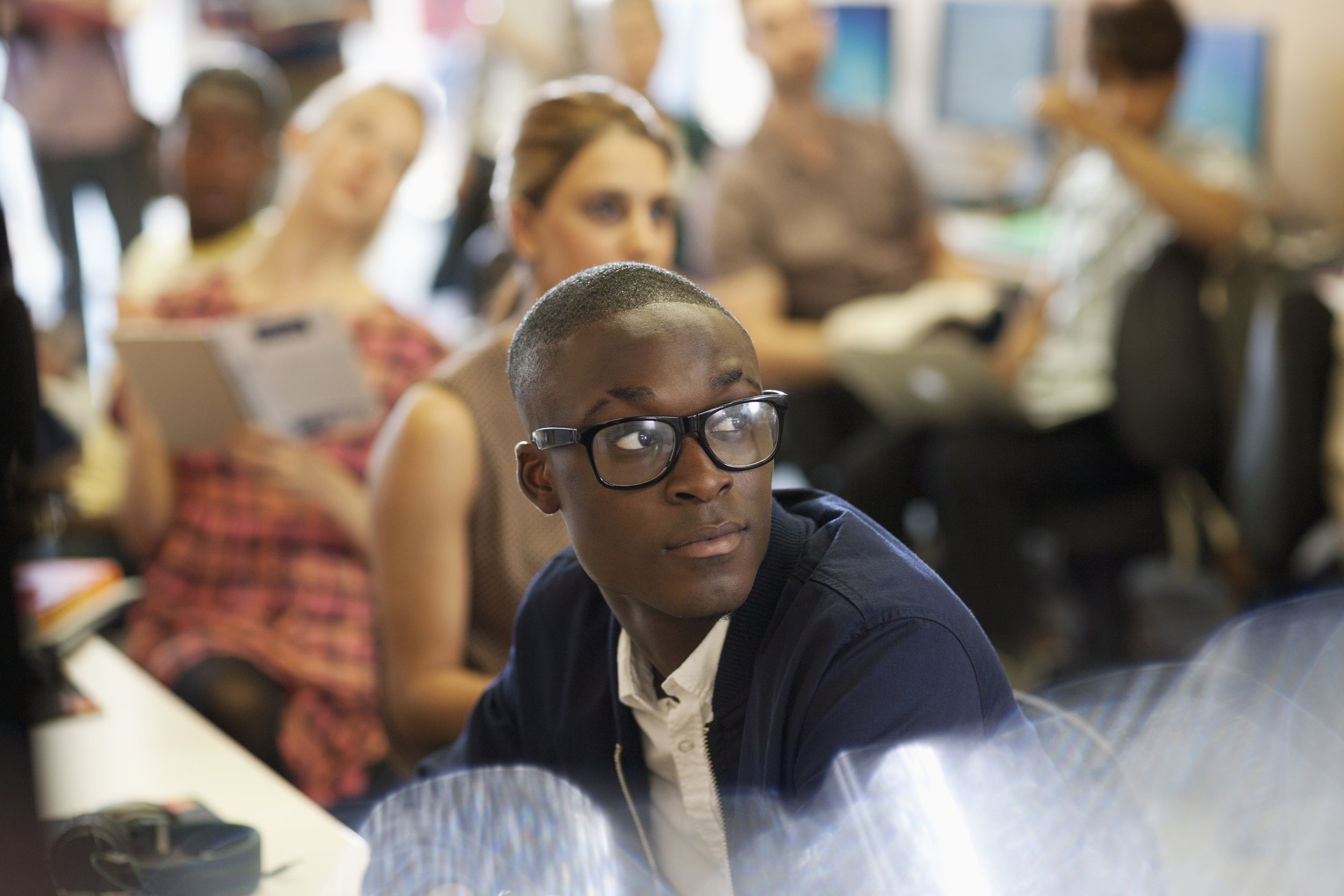 A young man wearing glasses and a dark jacket looks to the side in a busy classroom or lecture hall, with other students in the background reading and paying attention.