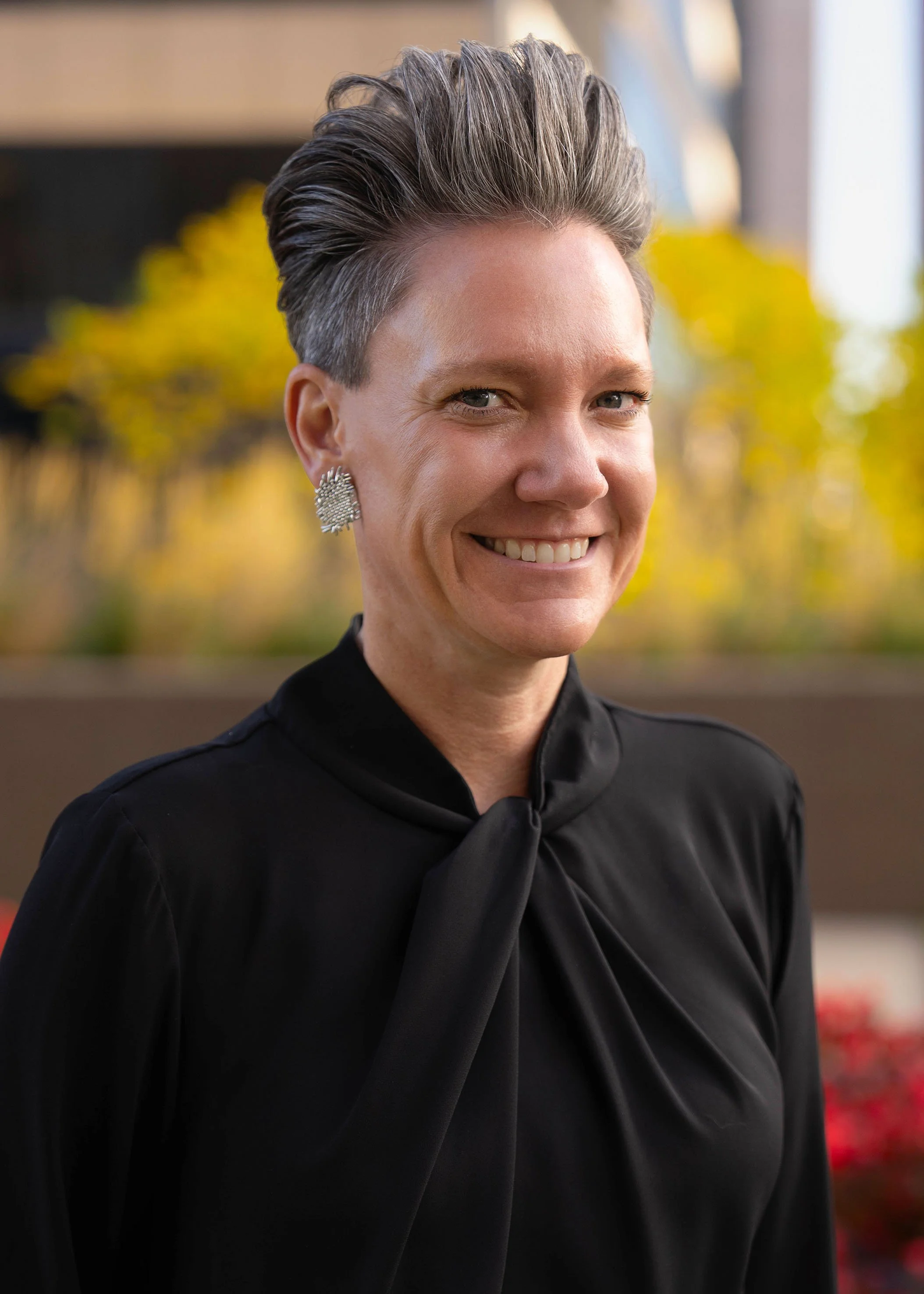 A woman with short, gray hair, wearing large earrings and a black top with a bow at the neck, smiling outdoors in front of blurred trees with fall foliage.