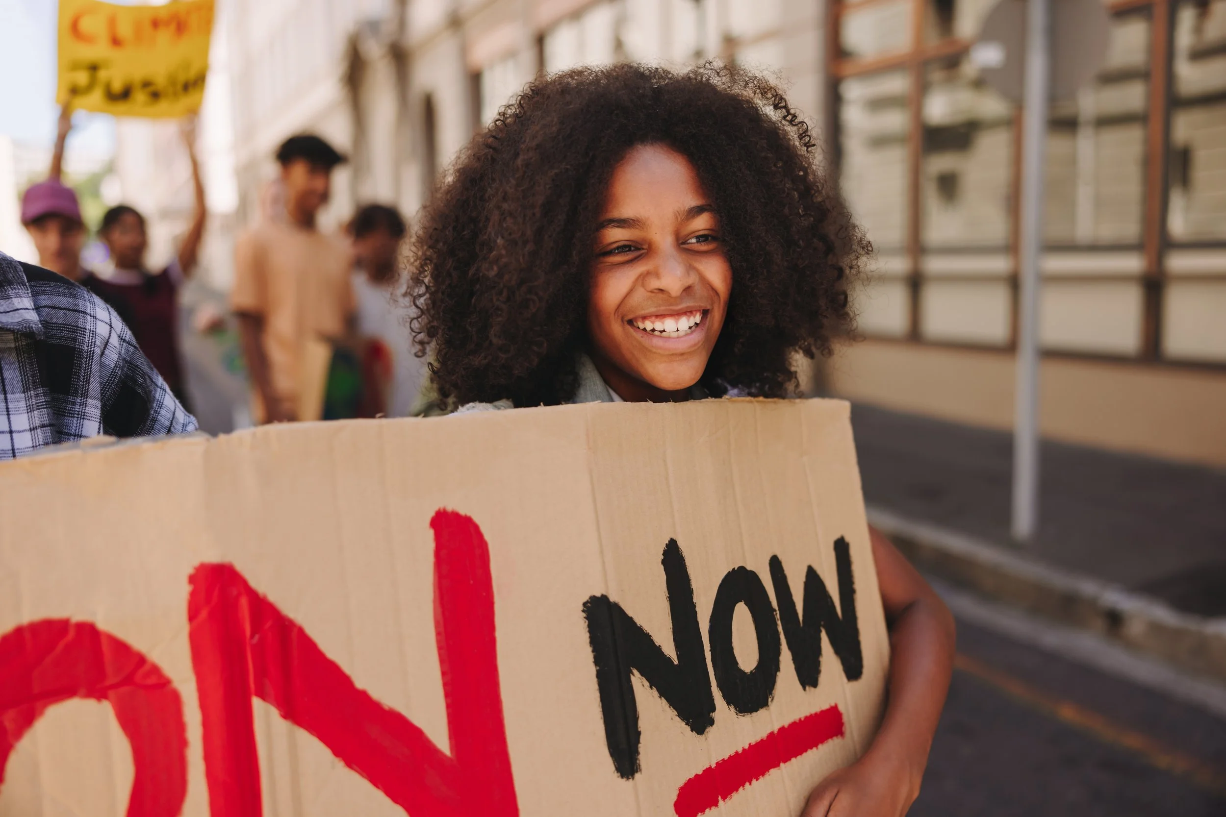 Smiling woman holding a protest sign that says 'NOW' on a city street during a demonstration with other protesters in the background.