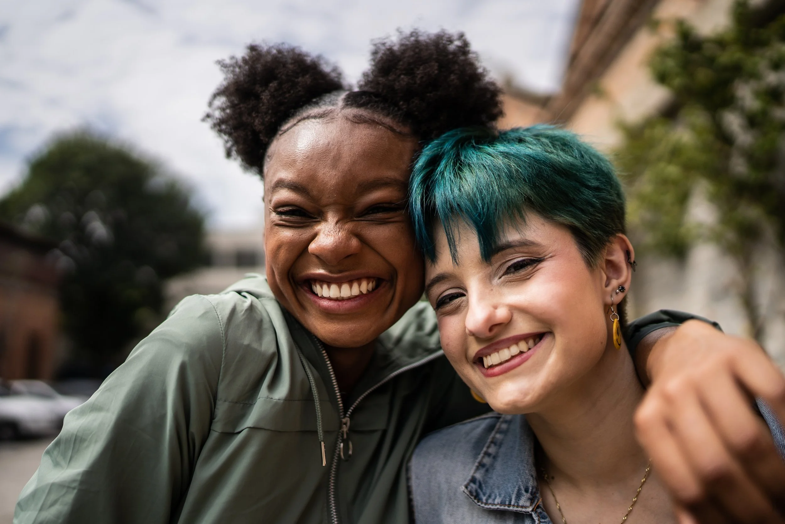 Two women smiling and hugging outdoors, one with natural afro hairstyle and the other with short colorful teal hair.