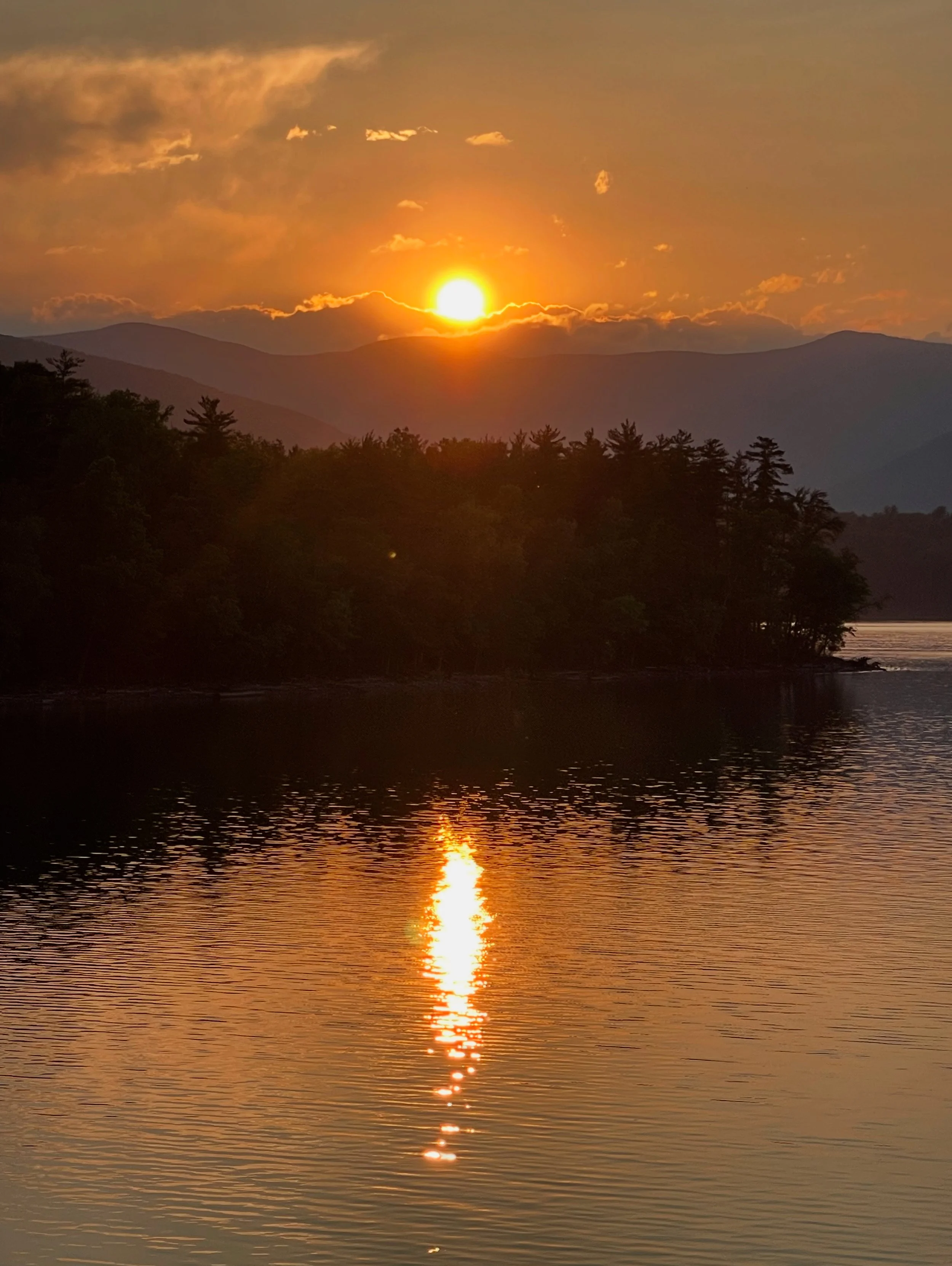 Sunset over a mountain range with a reflection in a calm lake below, surrounded by trees.