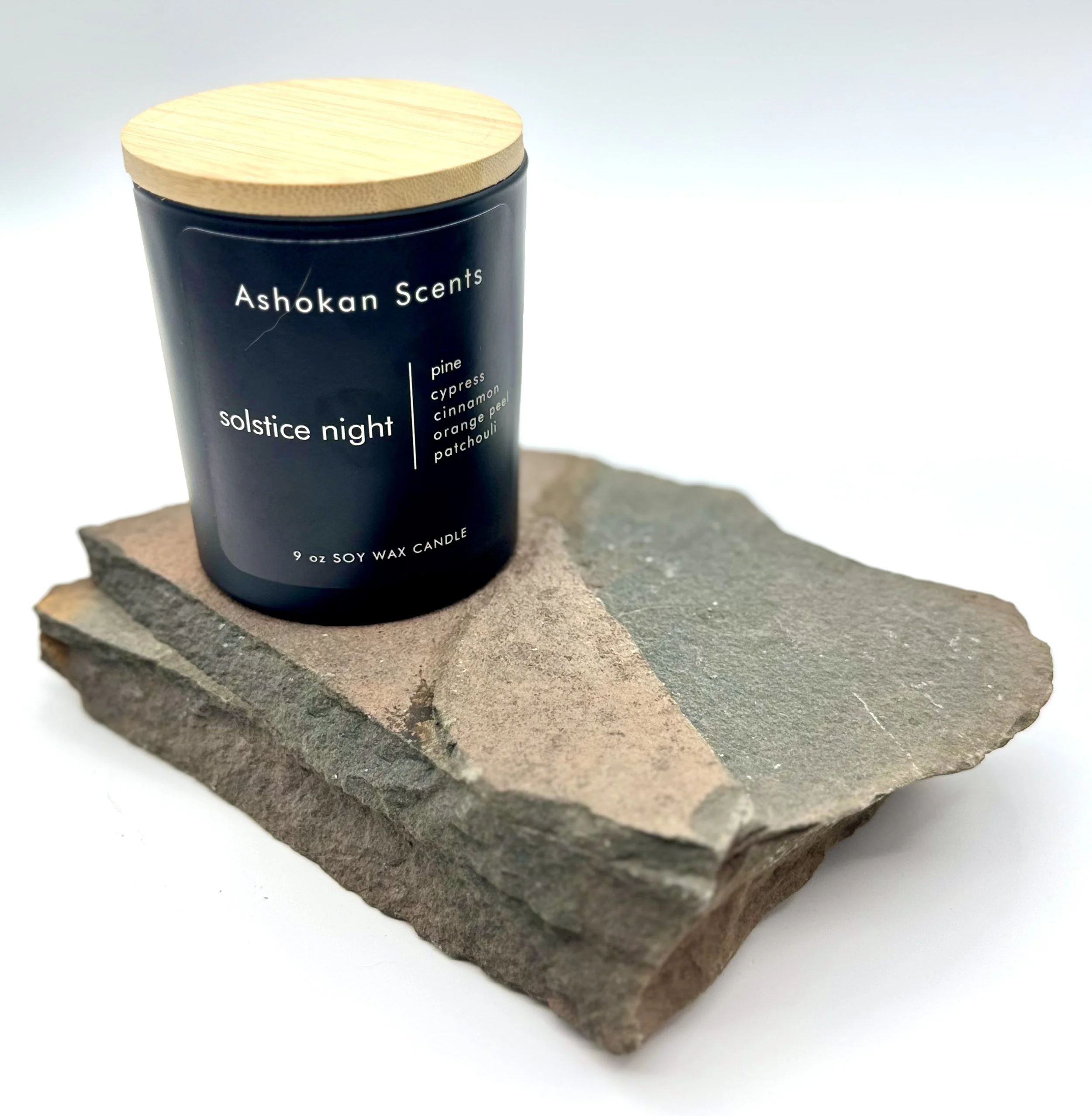 A black candle jar with a wooden lid labeled "Ashokan Scents" and "solstice night," placed on a piece of natural stone.